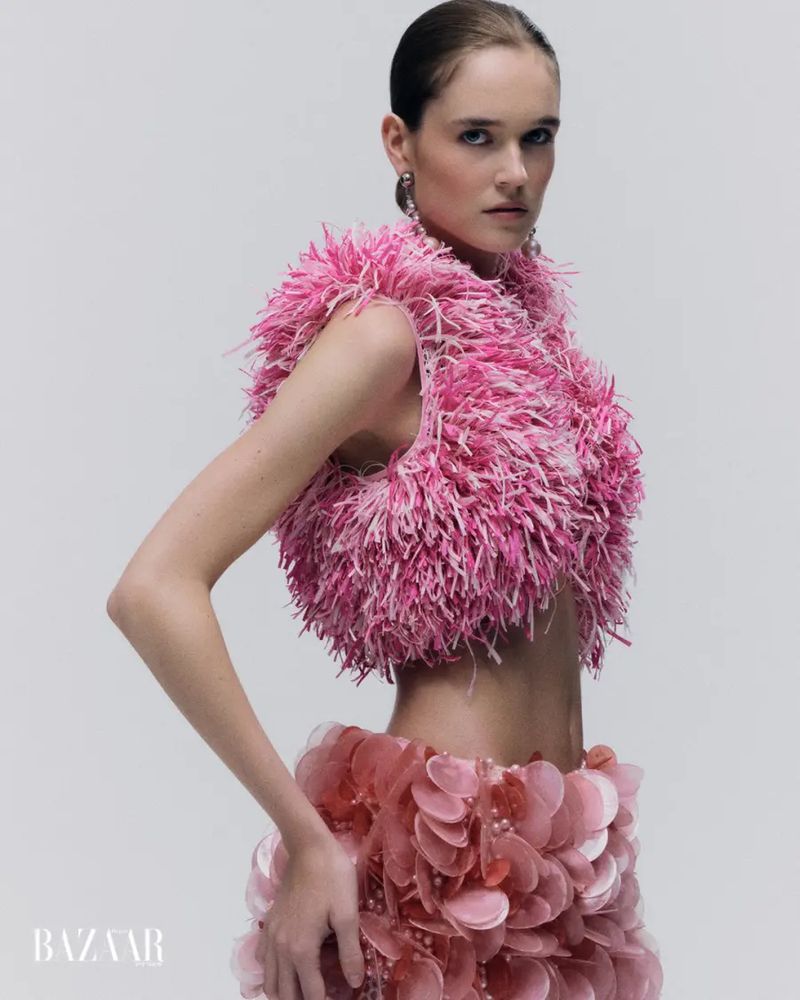 Model wearing a textured pink fringe crop top and circular sequin skirt posing against a light grey studio background for Harper's Bazaar.
