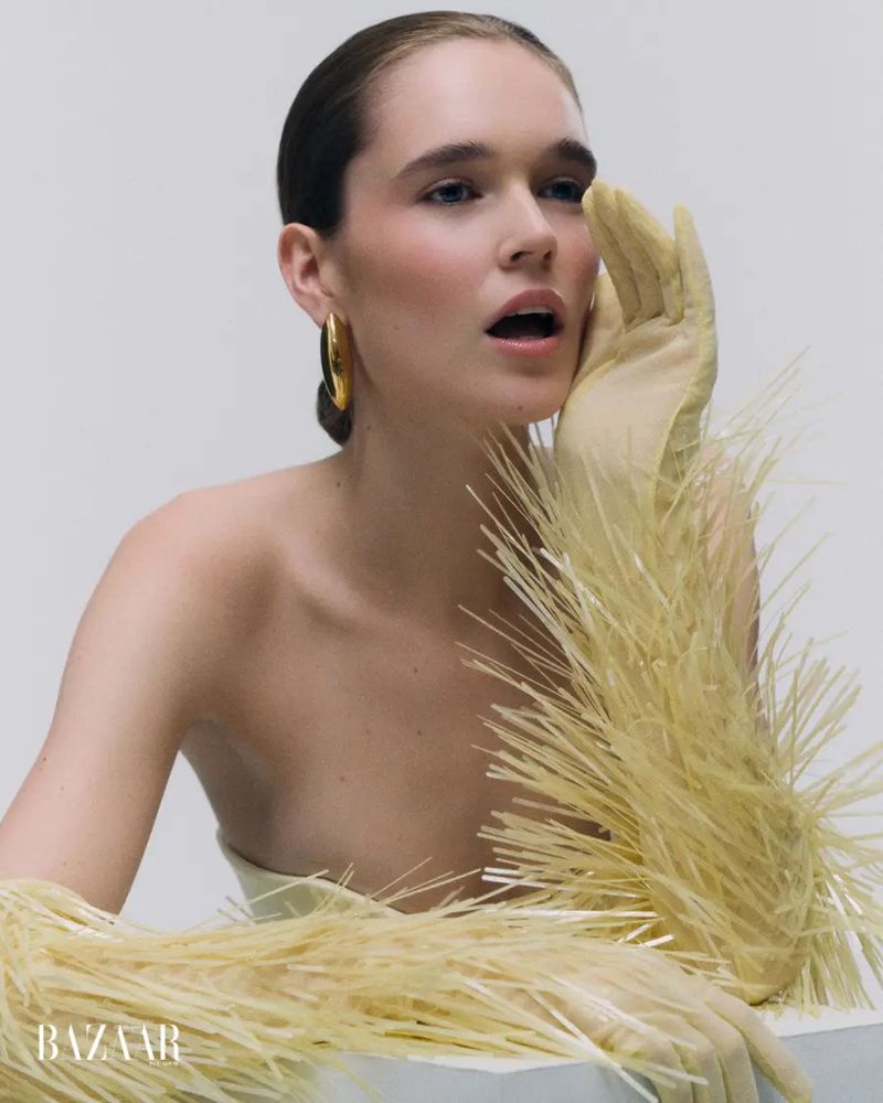 Model wearing textured straw-like fringed yellow gloves and gold earring posing with hand to face for Harper's Bazaar Ecuador.