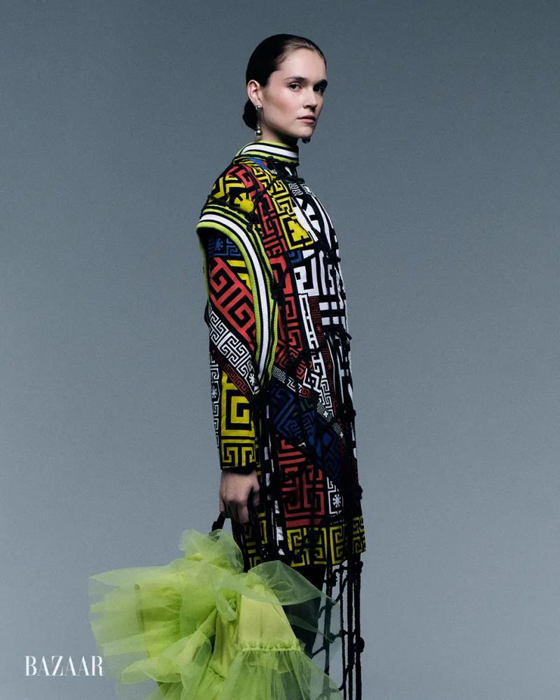 Model posing in a multicolor geometric patterned coat holding a bright lime green tulle accessory against a grey backdrop.
