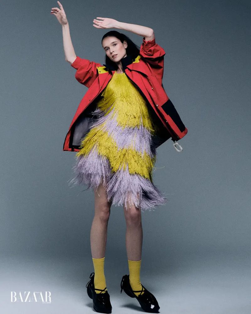 Model posing in a yellow and lavender tiered fringe dress with an oversized red windbreaker jacket against a grey background.