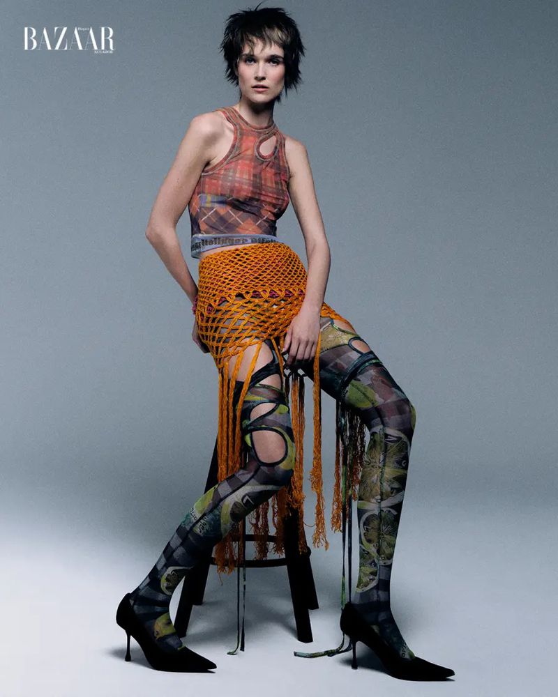 Model wearing an orange fringe macrame skirt, printed crop top, and patterned cutout tights seated on a stool in a studio.