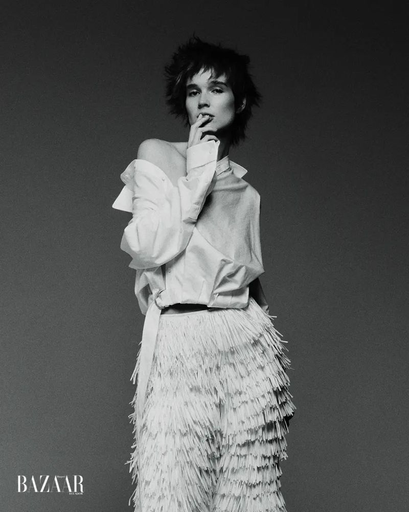 Black and white portrait of a model wearing an off-shoulder white shirt and white tiered fringe pants in a studio.