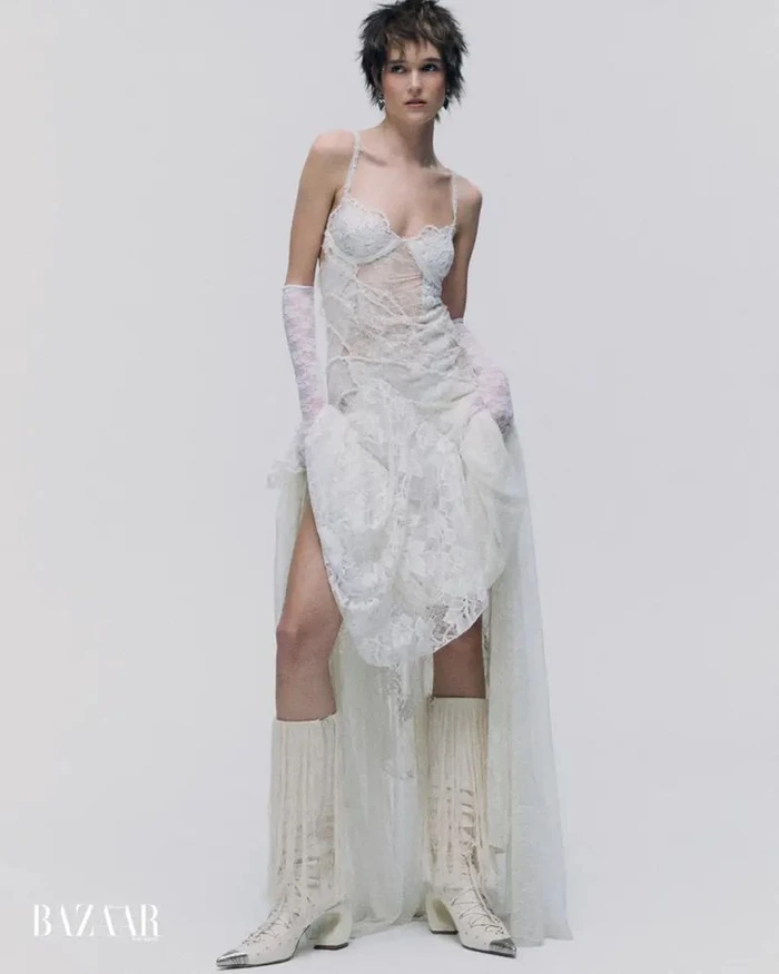 Model with short dark hair wearing a sheer white lace gown, lace gloves, and white fringe boots against a seamless studio background.
