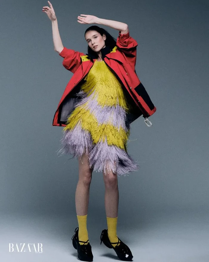 Model posing in a yellow and lavender tiered fringe dress with an oversized red windbreaker jacket against a grey background.