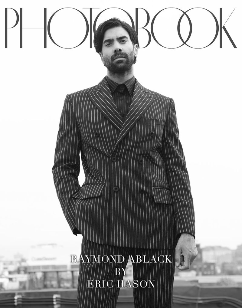 Black and white portrait of a bearded man wearing a pinstripe suit. Outdoor rooftop setting with cityscape. Fashion editorial photography.