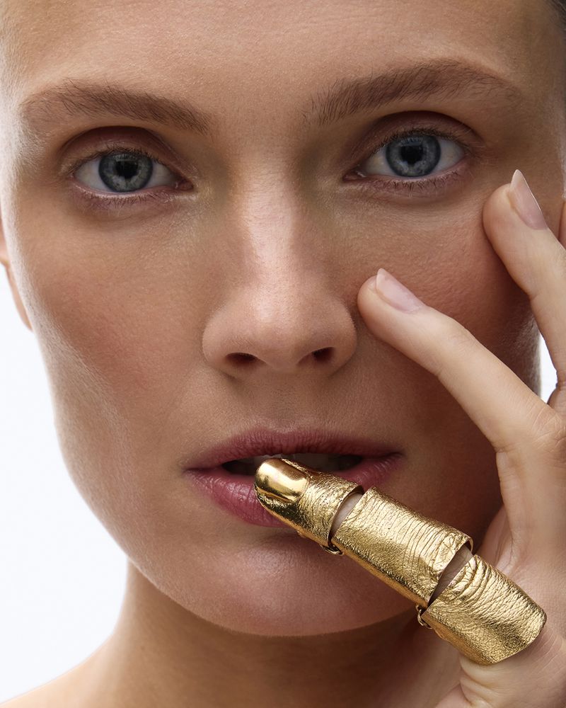 Close-up portrait of model Constance Jablonski touching her lip with a textured gold full-finger ring against a white studio background.