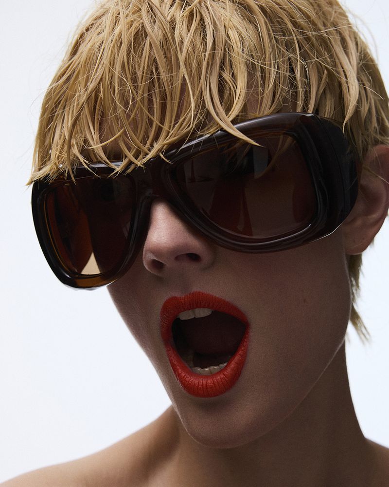 Constance Jablonski wearing large dark sunglasses and red lipstick against a bright white background.