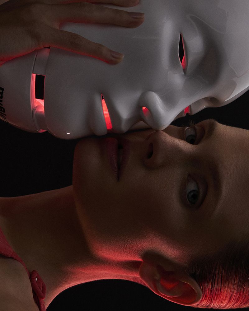 Constance Jablonski posed closely beside an illuminated red LED light therapy mask against a dark studio backdrop.