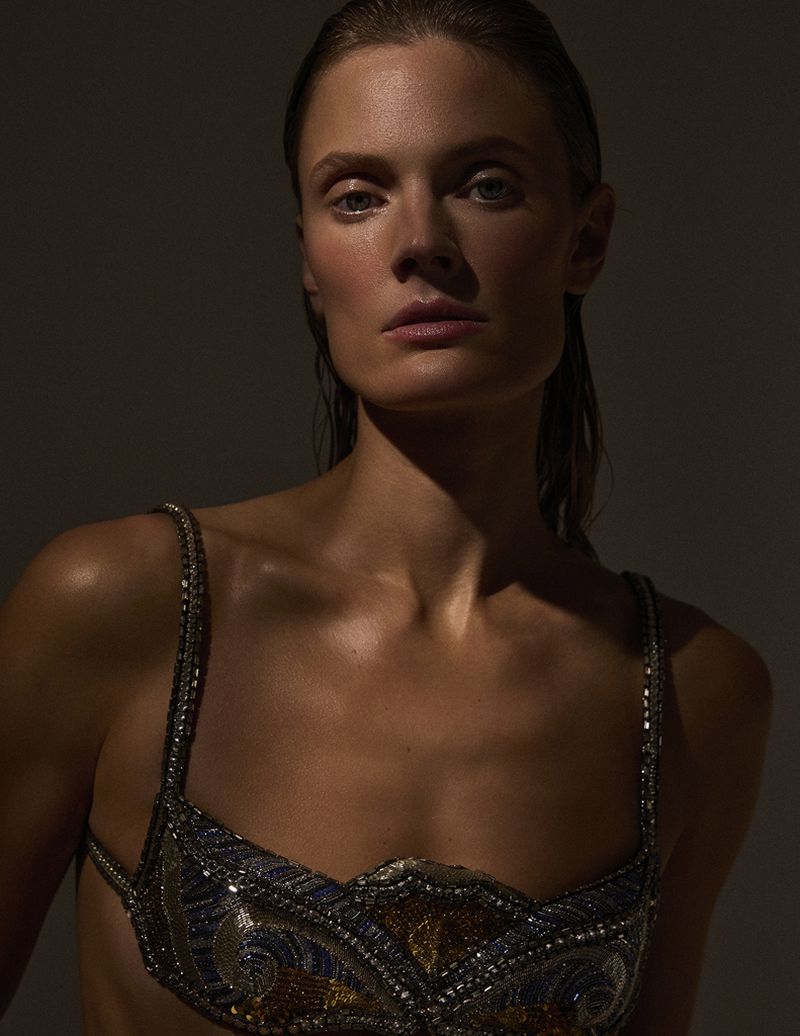Constance Jablonski wearing a beaded metallic bralette with slicked-back hair against a dark studio background.