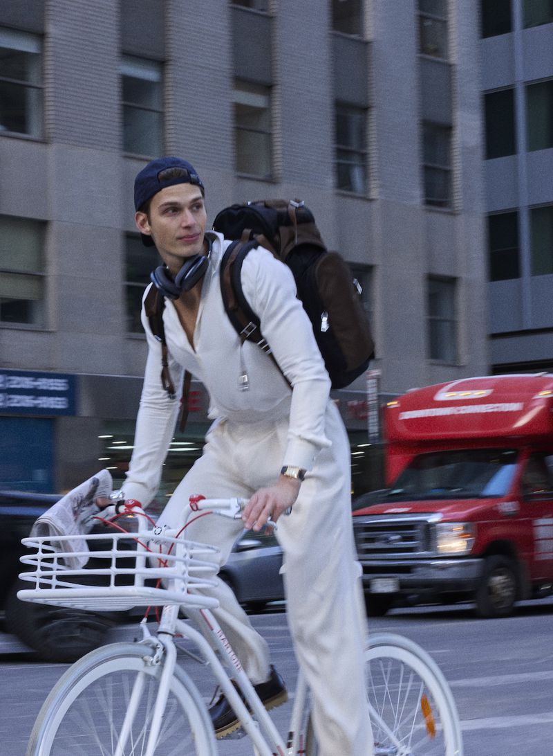 Model Astrid Voss rides a white bicycle through city streets wearing an all-white ensemble, backwards cap, and backpack.