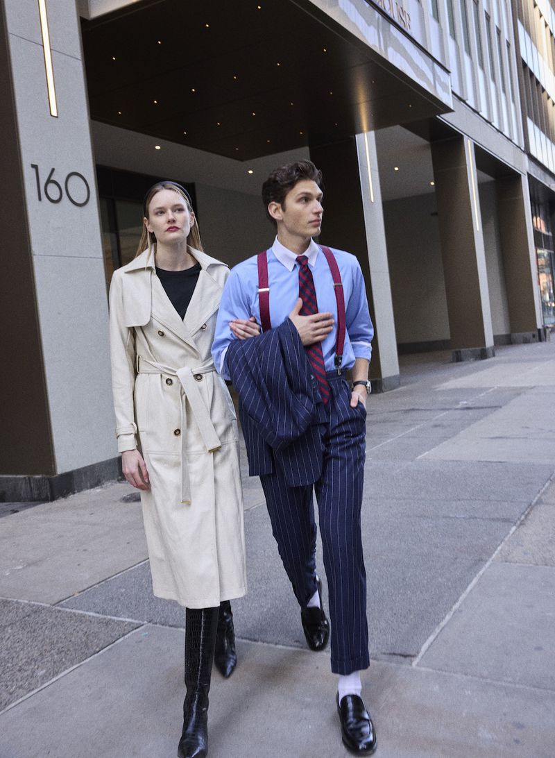 Astrid Voss and a male model walking arm-in-arm on a city sidewalk wearing a cream trench coat and navy pinstripe suit.