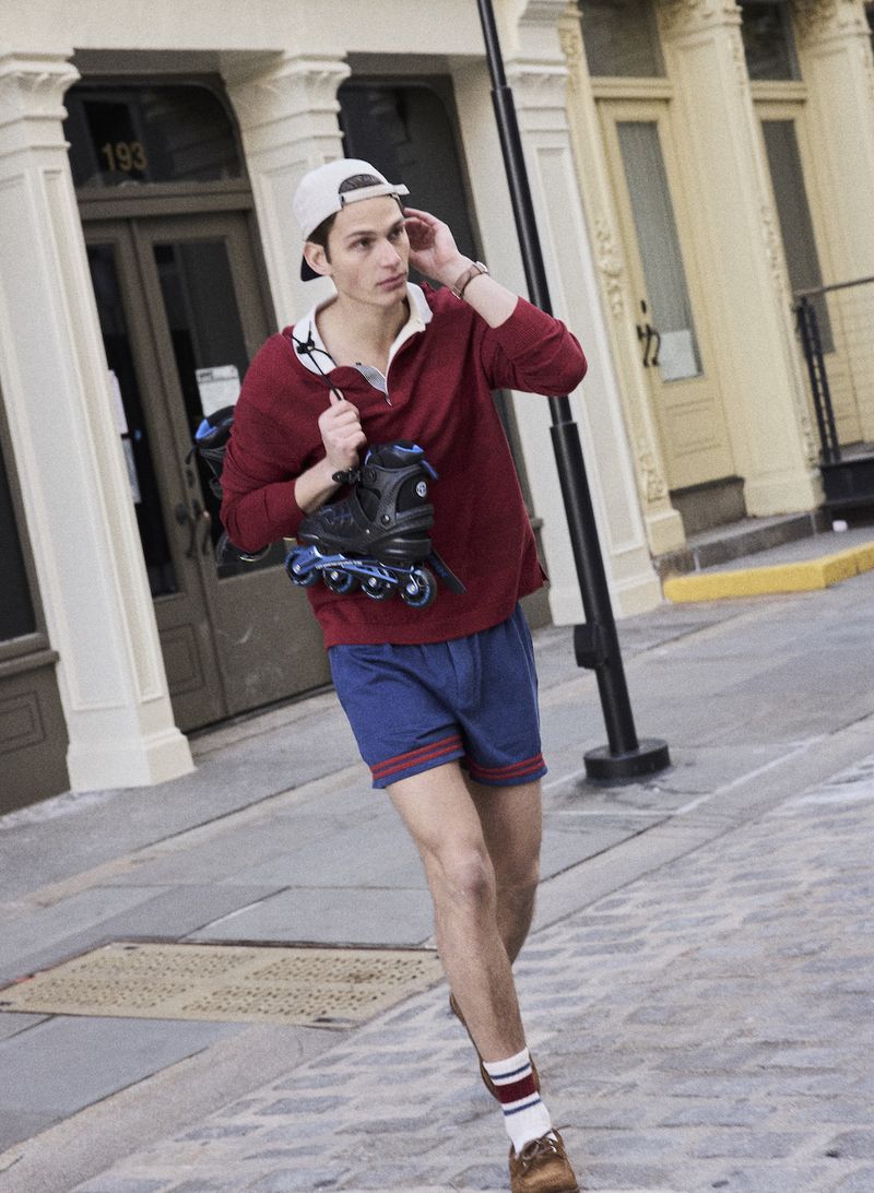A man walking on a cobblestone street wearing a burgundy long-sleeve polo, blue shorts, backwards cap, and carrying inline skates.