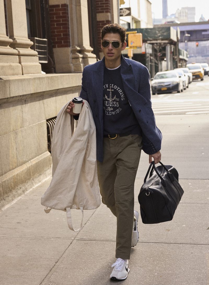 A man in sunglasses, blue blazer and olive trousers walks down a city sidewalk carrying a garment bag and duffel bag.