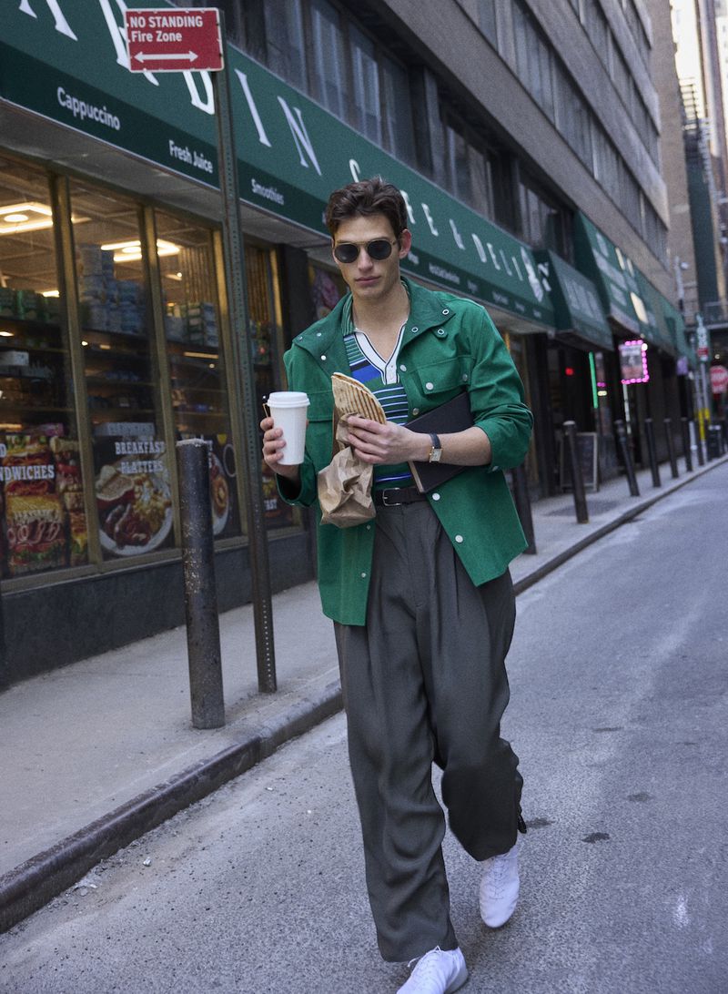 A man in a green jacket and sunglasses walking down a city street holding a coffee cup and a paper bag.