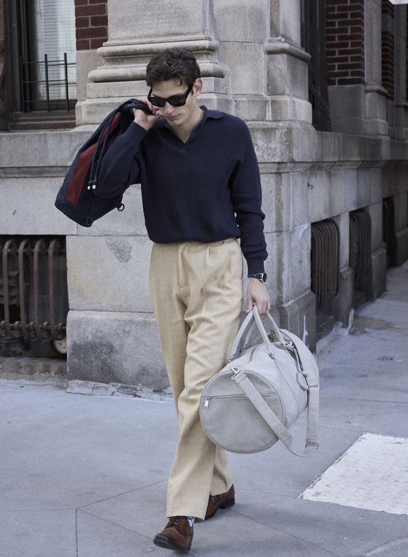 Man in a navy polo sweater, beige pleated trousers, and sunglasses walking with a grey duffle bag outside a stone building.