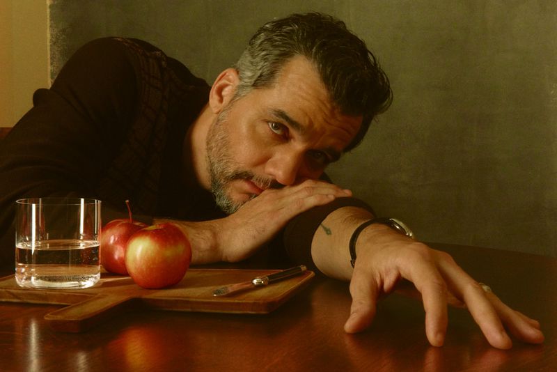 Actor Wagner Moura resting his head on a wooden table beside two red apples and a glass of water.