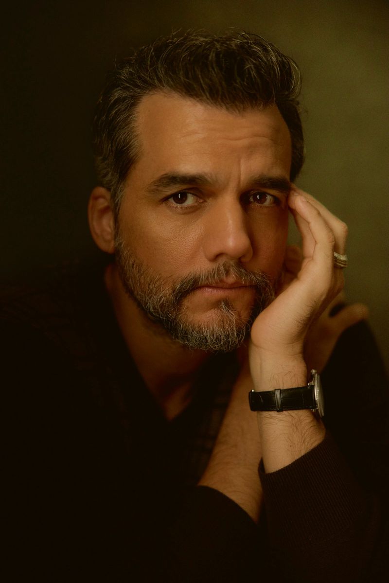 Portrait of a middle-aged Caucasian man with salt and pepper hair, a beard, wearing a black shirt and wristwatch, set against a dark background.
