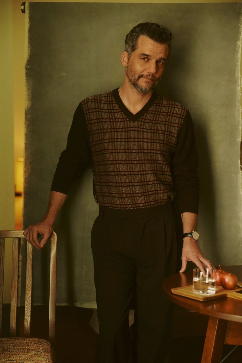Wagner Moura standing between a wooden chair and table wearing a patterned knit sweater and dark trousers.