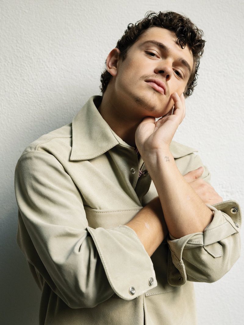 Noah Jupe posing in a beige button-up overshirt with his hand under his chin against a textured off-white wall.