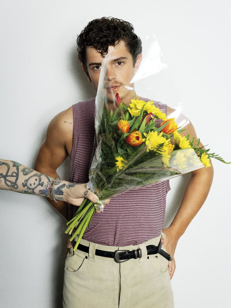 Noah Jupe in a striped tank top and beige trousers posing behind a flower bouquet held by a tattooed arm against a white wall.