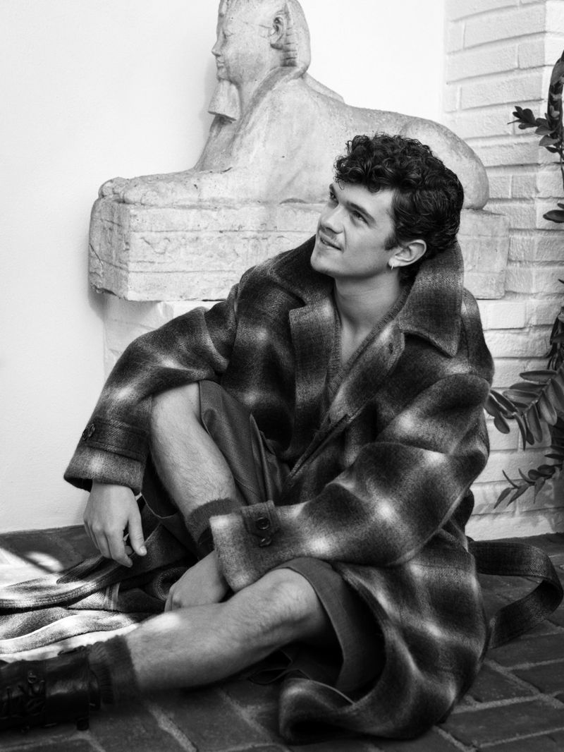 Actor Noah Jupe seated on a brick ground in an oversized plaid coat leaning against a stone sphinx statue in black and white.