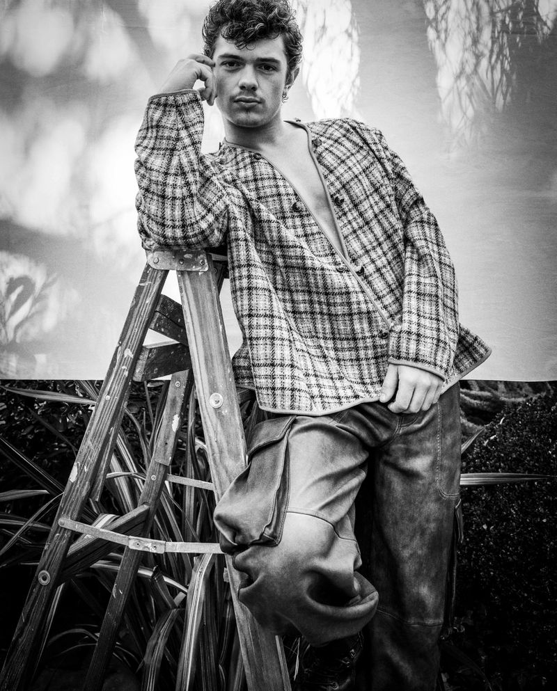 Monochrome portrait of Noah Jupe leaning on a weathered ladder wearing an open plaid jacket and cargo pants outdoors.