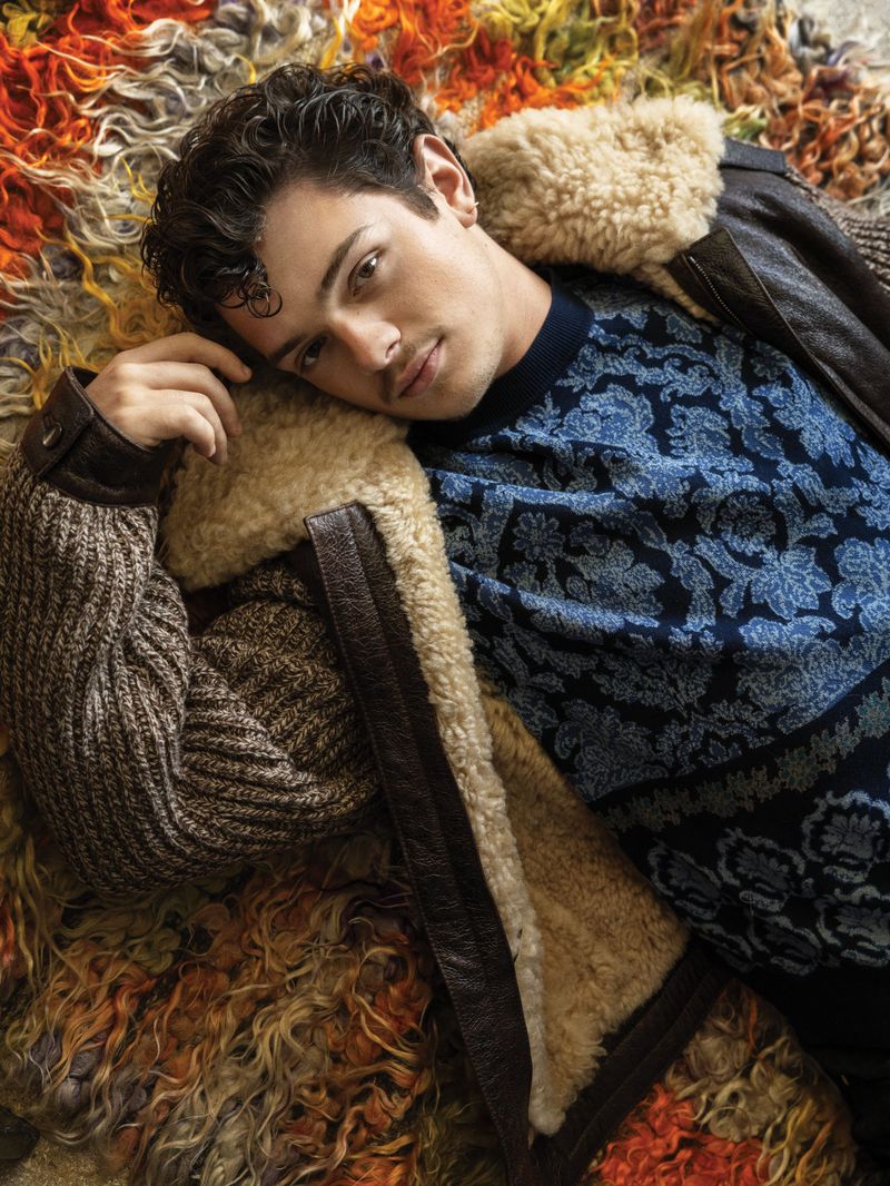 Noah Jupe reclining on a colorful textured shag rug wearing a blue patterned knit sweater and a shearling-lined brown jacket.