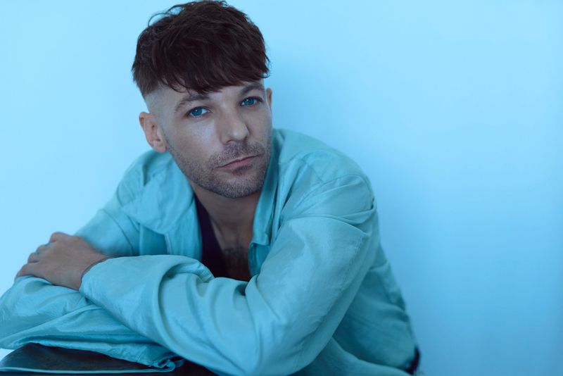 Louis Tomlinson leans forward in a light blue jacket against a pale blue background in an editorial studio portrait.