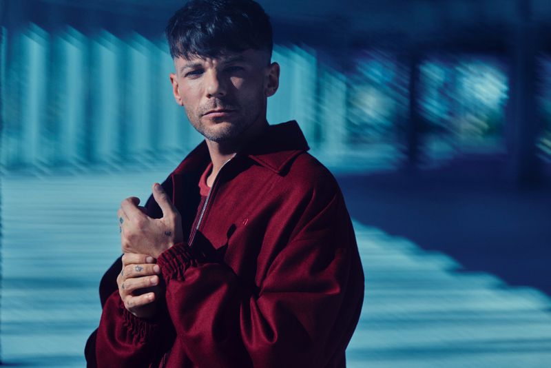 Louis Tomlinson wearing a burgundy jacket and looking directly at the camera with hands clasped against a blurred blue background.