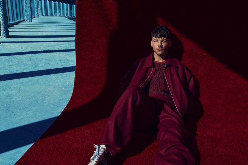 Musician Louis Tomlinson reclining on a curved red carpet set dressed in a matching maroon tracksuit and sneakers.