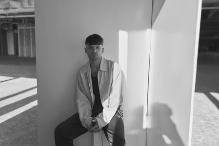 Louis Tomlinson seated against a white panel in a black and white editorial portrait taken in an industrial space.