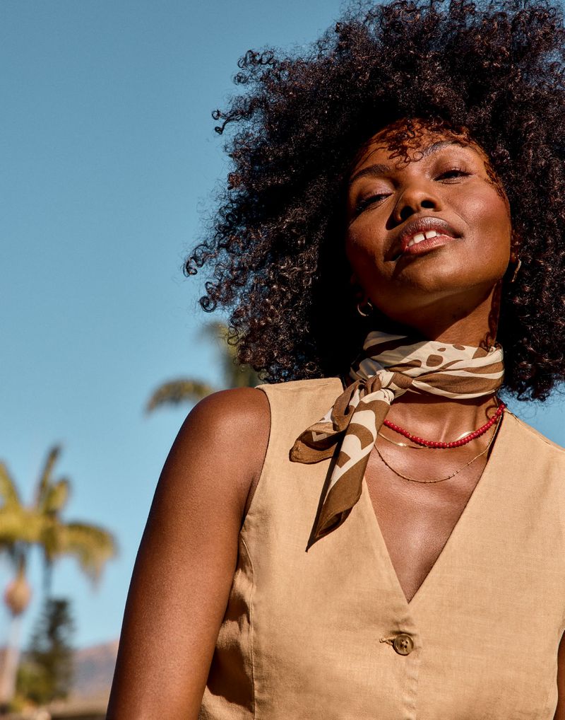 Black woman with curly hair wearing a beige sleeveless vest, patterned neck scarf, and necklace against a clear blue sky.