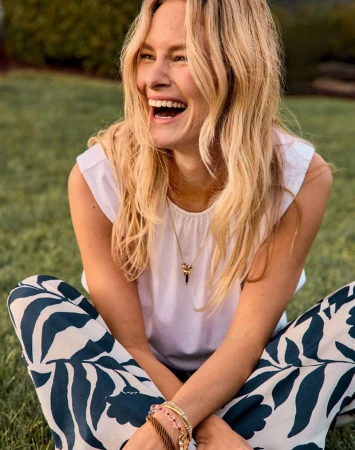 Blonde woman laughing while seated on grass wearing a white top and navy botanical print pants.