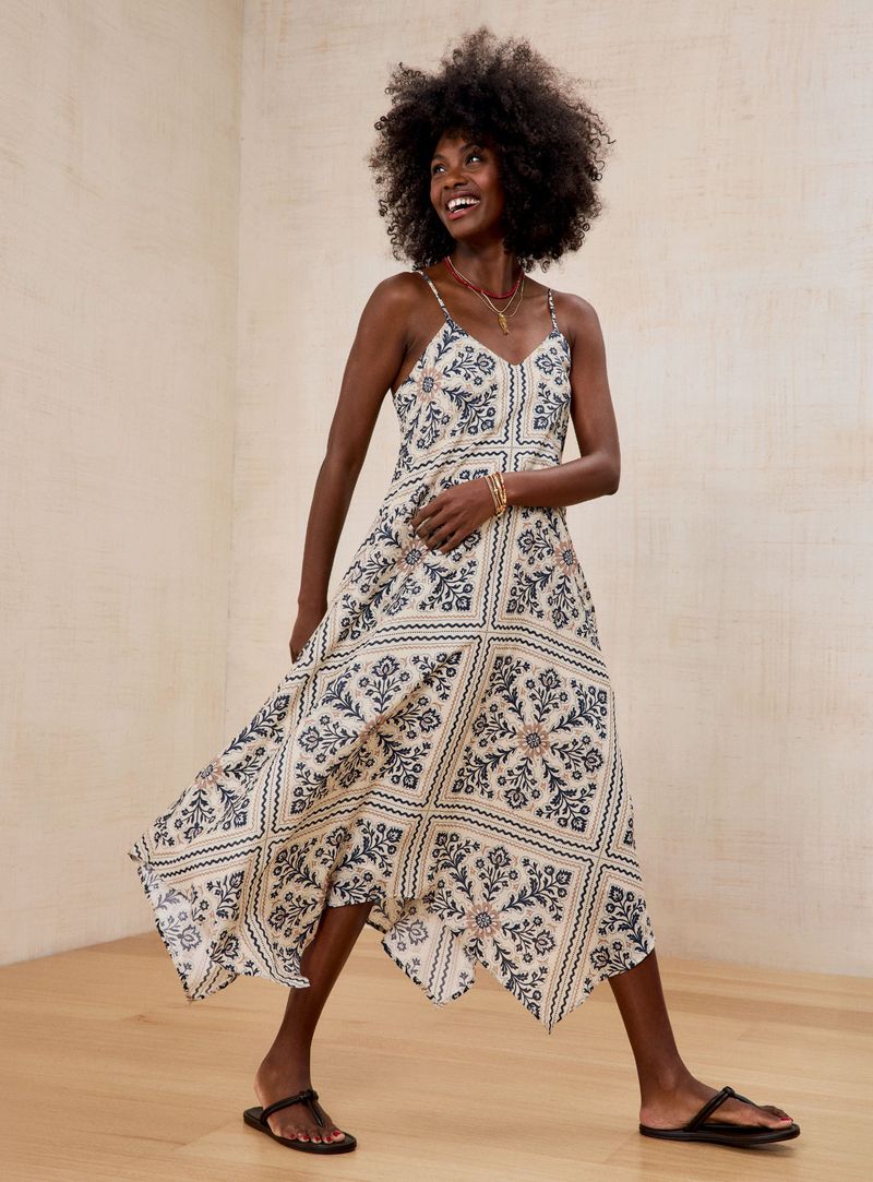 Black woman with afro hair smiling in a patterned handkerchief-hem sundress and flat black sandals in a studio setting.