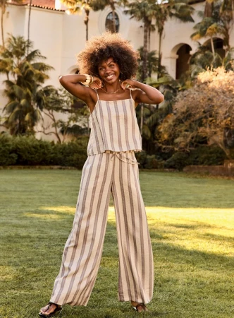 Smiling Black woman with voluminous curly hair wearing a striped linen tank and matching wide-leg trousers on a sunlit lawn.