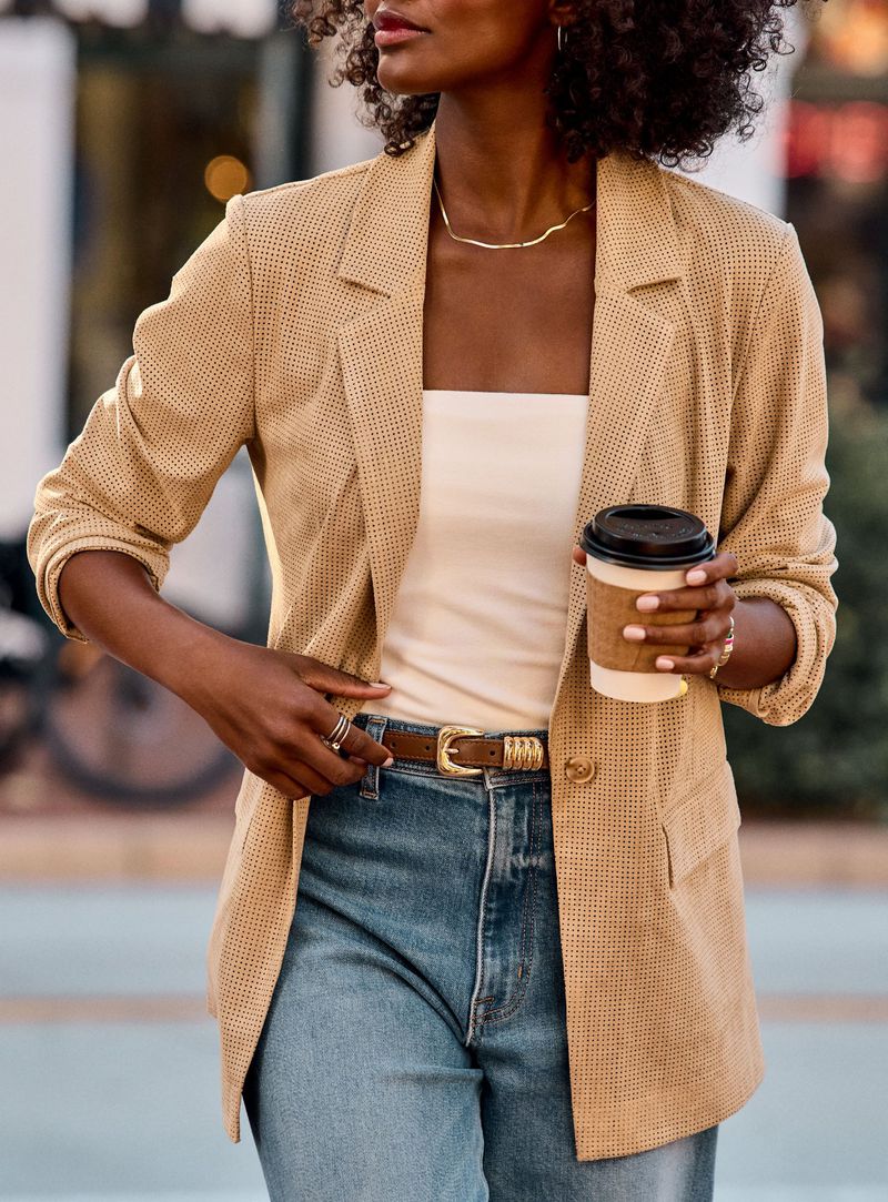 Woman wearing a textured tan blazer, white top, and blue jeans holding a paper coffee cup on a city sidewalk.
