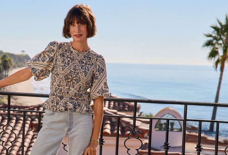 Woman in patterned puff-sleeve top and light denim leaning against a balcony railing overlooking the ocean.