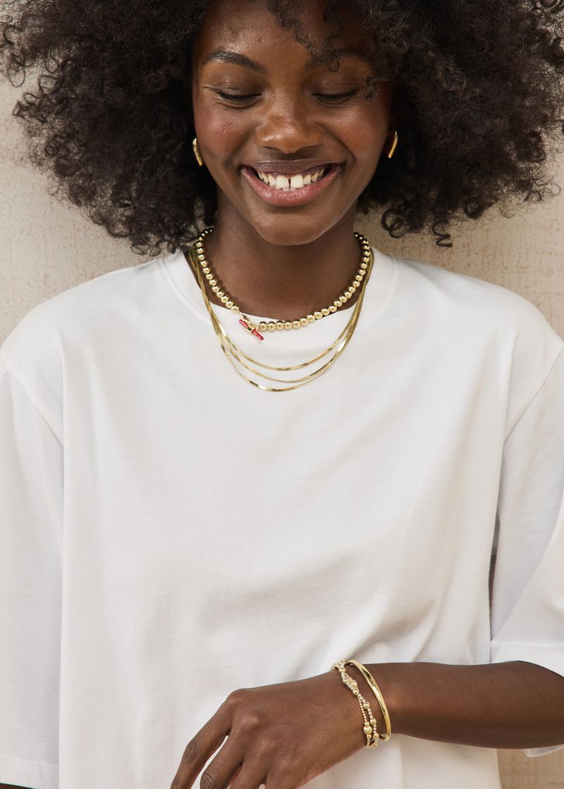 A smiling Black woman with curly hair looking down, wearing a white t-shirt and layered gold jewelry.