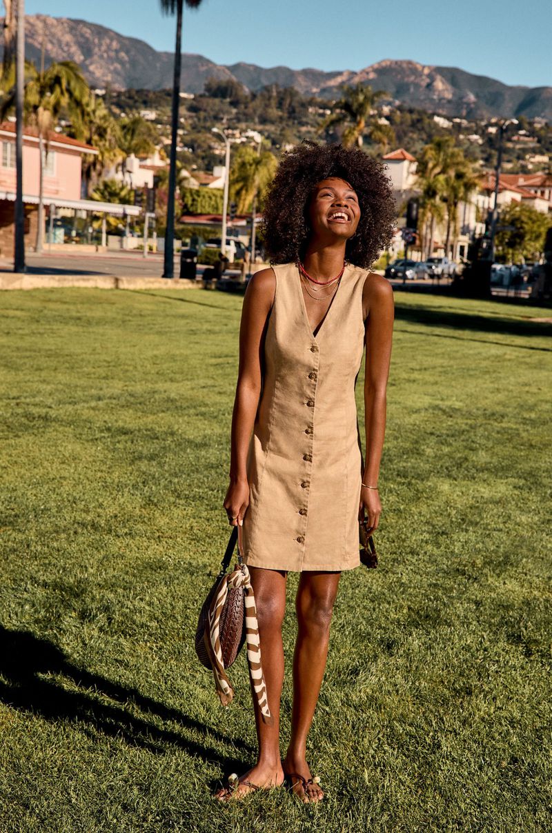 Smiling Black woman with voluminous curly hair wearing a beige button-front sleeveless dress and holding a handbag on a park lawn.
