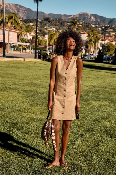 Smiling Black woman with voluminous curly hair wearing a beige button-front sleeveless dress and holding a handbag on a park lawn.