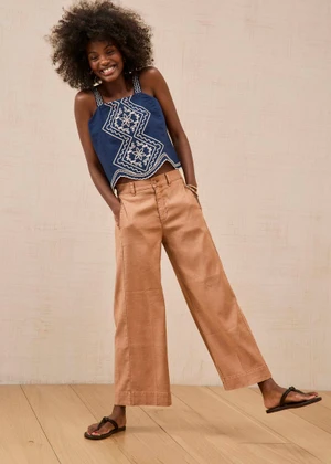 Smiling woman in a navy embroidered sleeveless top, tan wide-leg trousers, and black sandals posing in a studio.
