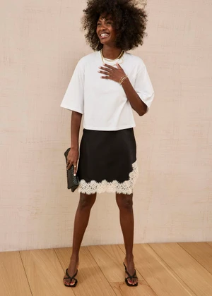 Black woman in a white boxy t-shirt and black lace-trimmed mini skirt smiling against a textured wall.