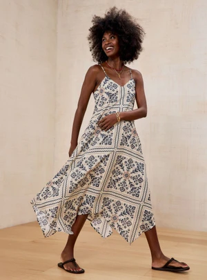 Black woman with afro hair smiling in a patterned handkerchief-hem sundress and flat black sandals in a studio setting.