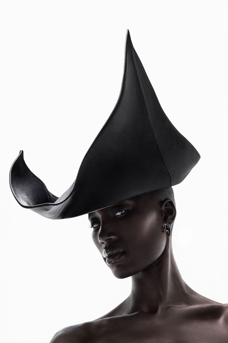 Close-up portrait of a Black woman wearing an avant-garde sculptural black hat against a bright white background.