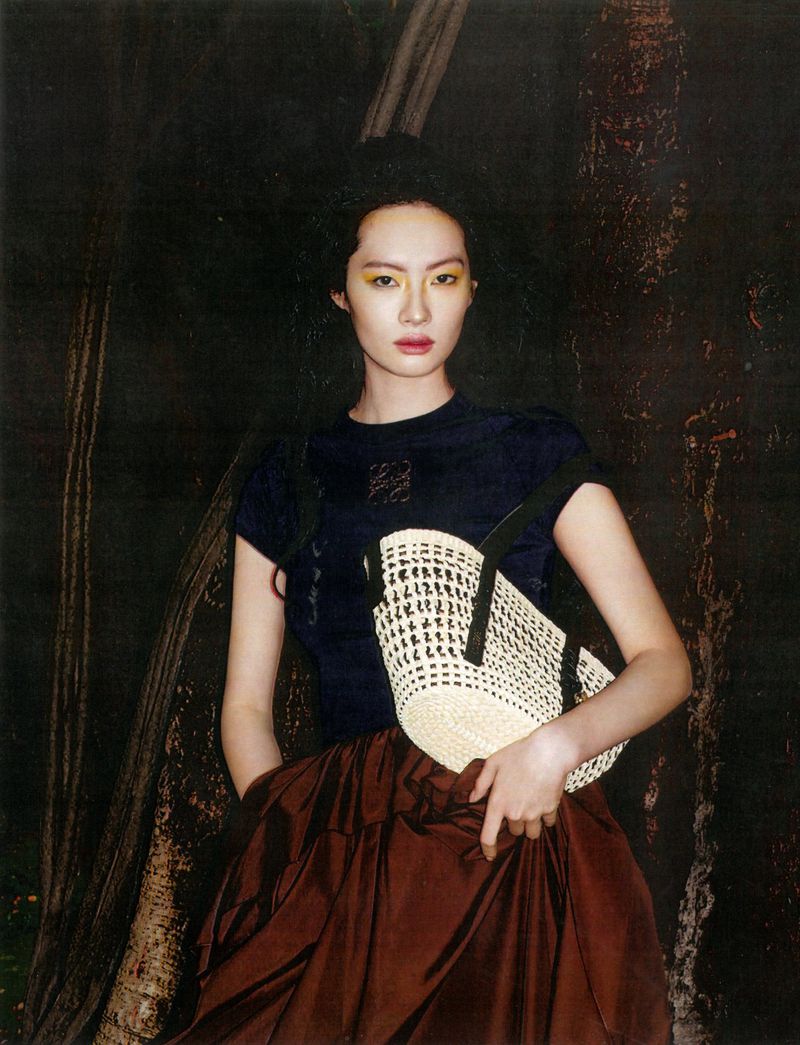 Model Wenyu Zhao in a navy top and voluminous brown skirt holding a cream woven bag against dark tree trunks.