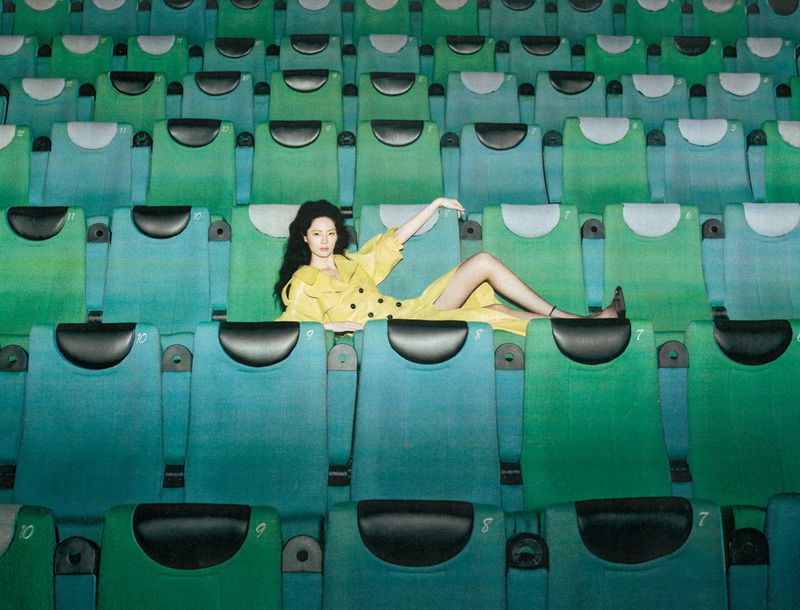 Model Wenyu Zhao wearing a bright yellow trench coat reclining across rows of green venue seats.