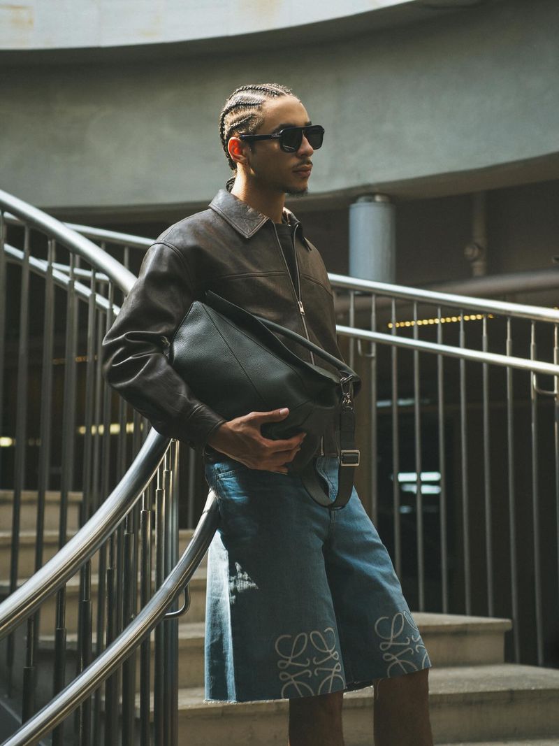Model Julez Smith wearing a brown leather jacket, sunglasses, and denim shorts while holding a black leather bag on a staircase.