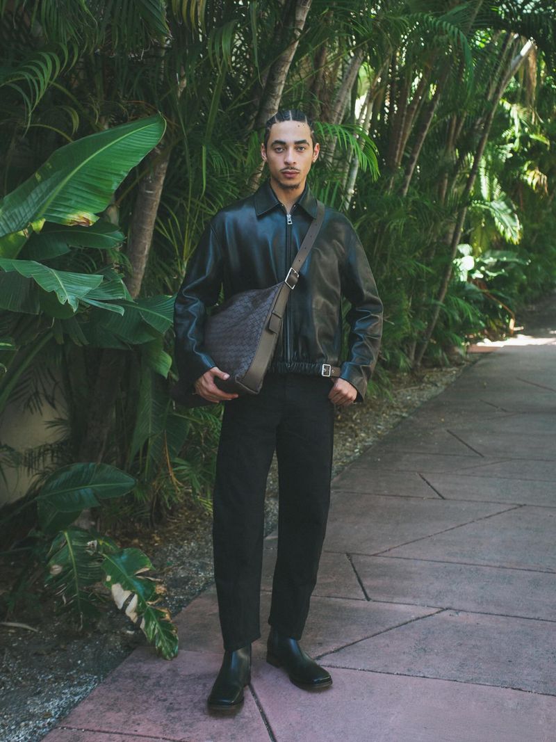 Model Julez Smith wearing a black leather jacket, trousers, and crossbody bag standing on a garden path lined with palm trees.