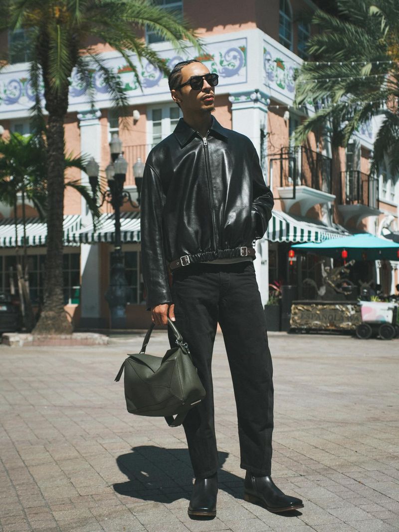 Julez Smith wearing a black leather jacket, dark sunglasses, and holding an olive green handbag in a sunlit plaza.