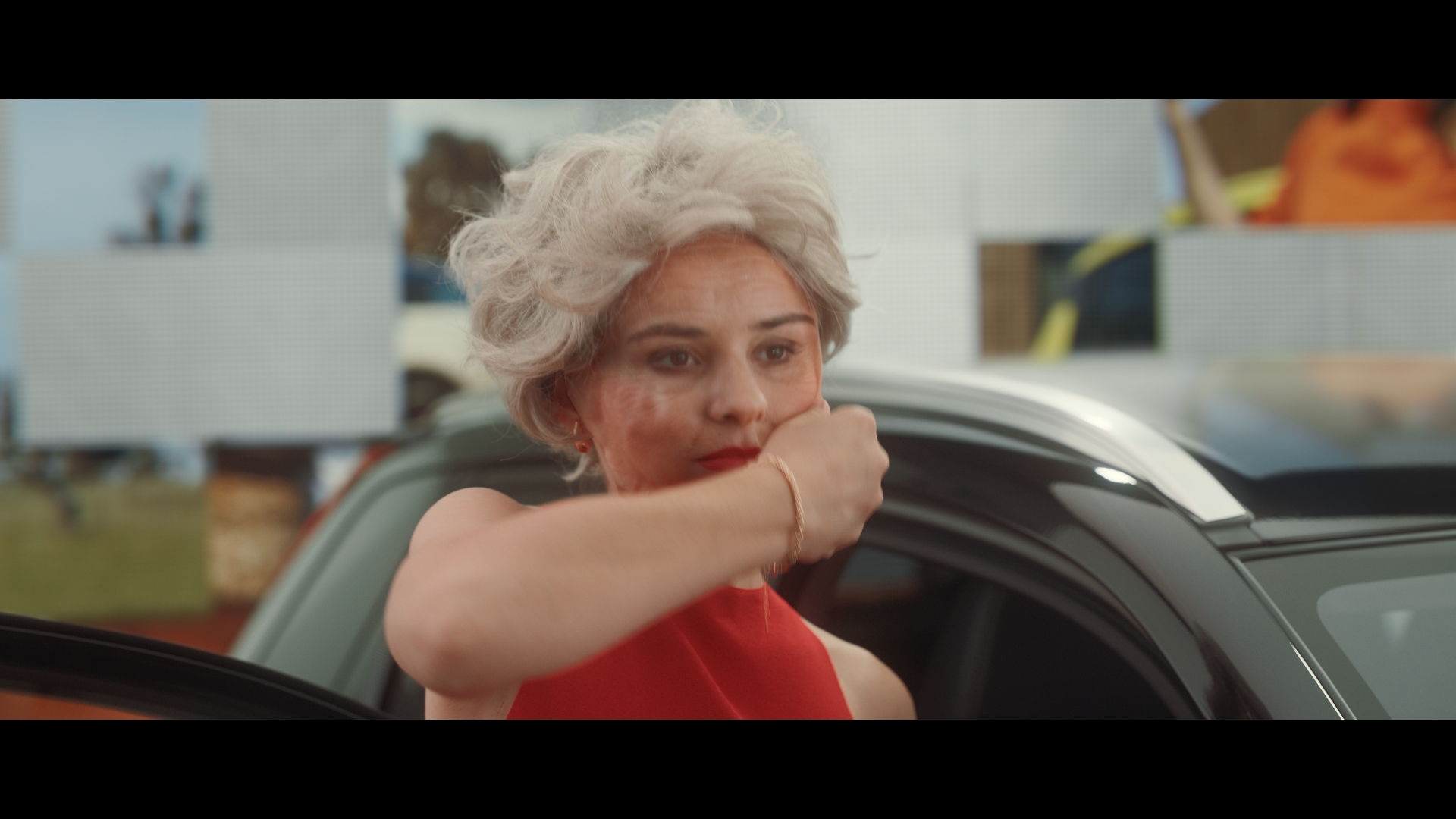 Woman with grey wig sitting in a car with a red top in a brightly lit interior. The woman is touching her face.